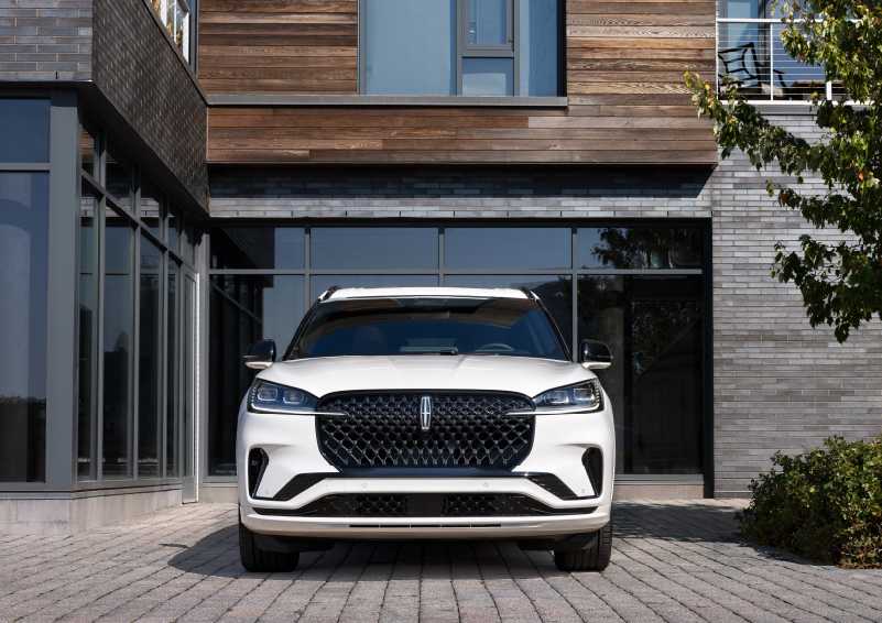 A white 2025 Lincoln Aviator® SUV is shown parked outside a modern building with gray brick and wood accents. | Ted Britt Chantilly Lincoln in Chantilly VA